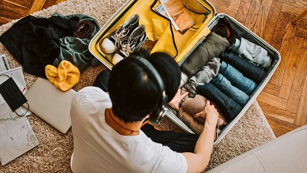 Overhead view of a person wearing headphones carefully packing a suitcase on a rug. The suitcase is filled with neatly rolled clothes and shoes, surrounded by travel essentials like a laptop and map, illustrating trip preparation and planning.